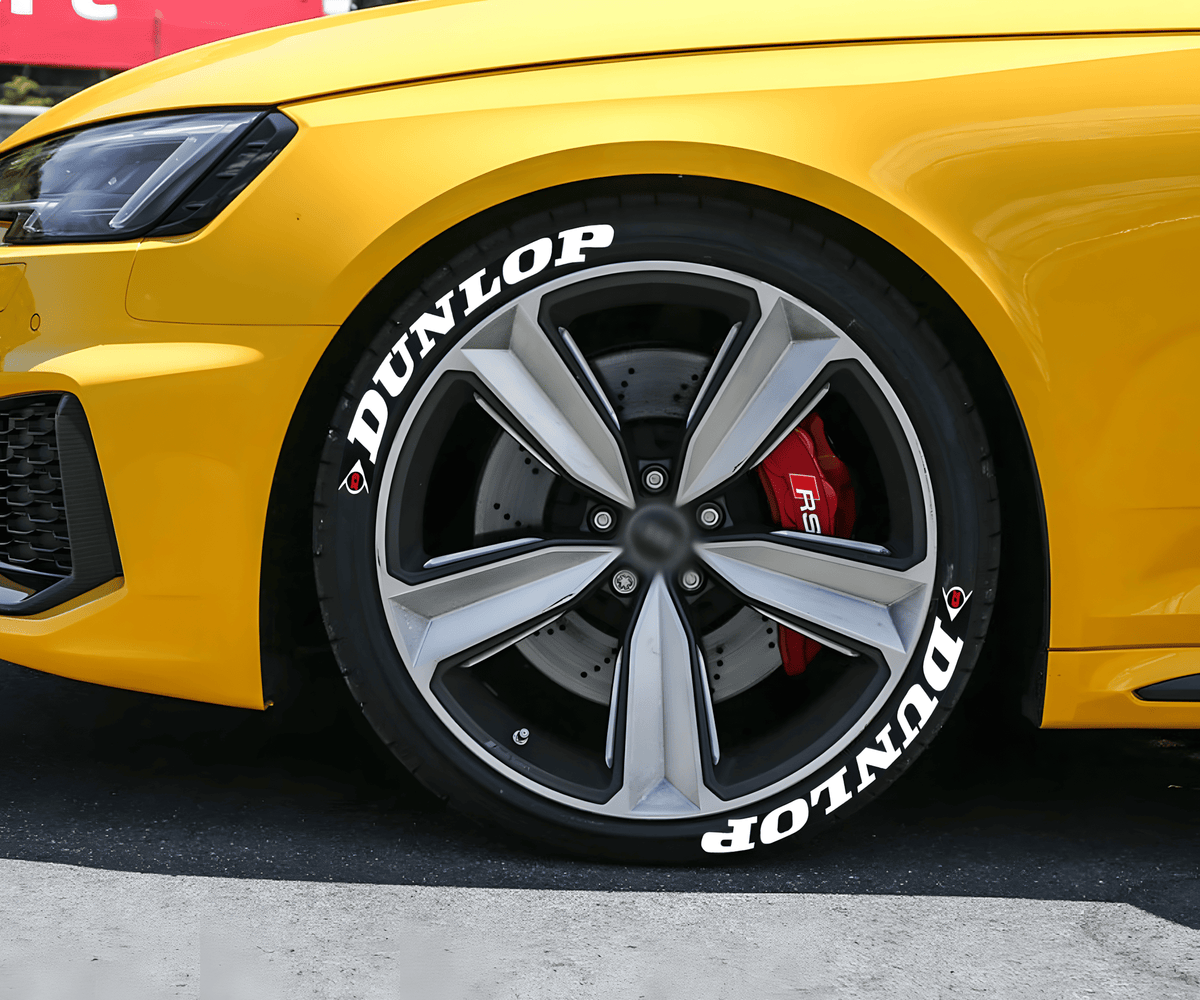 A yellow sports car featuring ChroMorpher Dunlop Performance Tire Lettering Decals in bold white text on black tires. The stylish and durable lettering enhances the vehicle’s racing-inspired look, adding a sleek and professional finish.