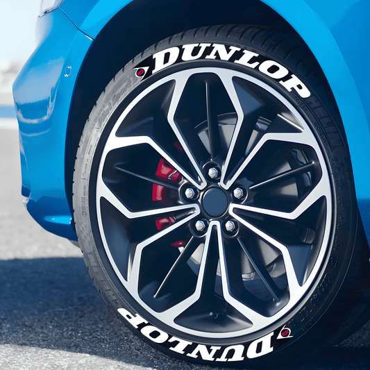 A close-up of a blue sports car wheel featuring ChroMorpher Dunlop Performance Tire Lettering Decals. The bold white "DUNLOP" text with a stylish red and black logo enhances the sporty aesthetic of the tire, giving it a professional racing look.