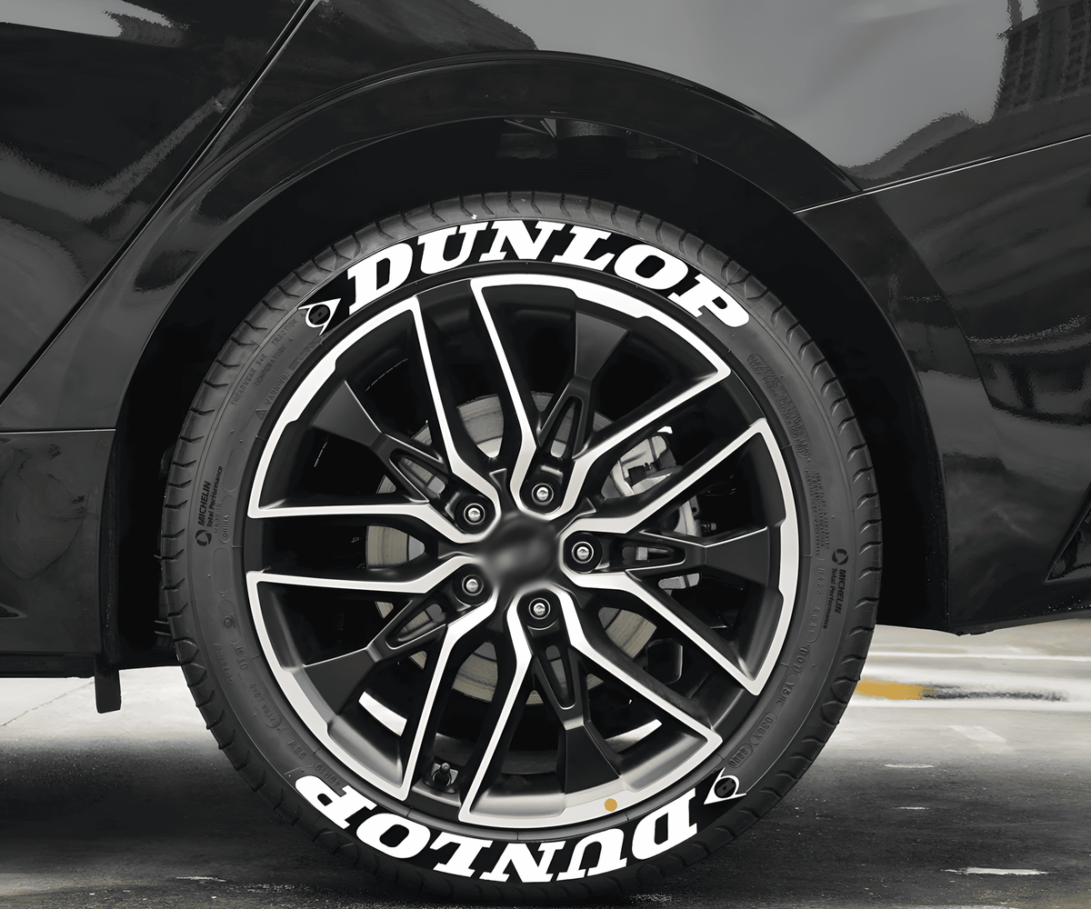 A black sports car fitted with ChroMorpher Dunlop Racing Tire Lettering Decals. The bold white "DUNLOP" text with a sleek racing-inspired design enhances the tire’s sporty look, perfect for high-performance vehicles.