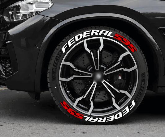 A black sports car with a close-up view of its front wheel featuring ChroMorpher FEDERAL 595 3D tire lettering decals in bold white and red. The decals enhance the tire's appearance, creating a racing-inspired look. The wheel has a modern, multi-spoke black and silver alloy rim with a high-performance braking system visible behind it. The car is parked on a concrete surface, with a blurred background.