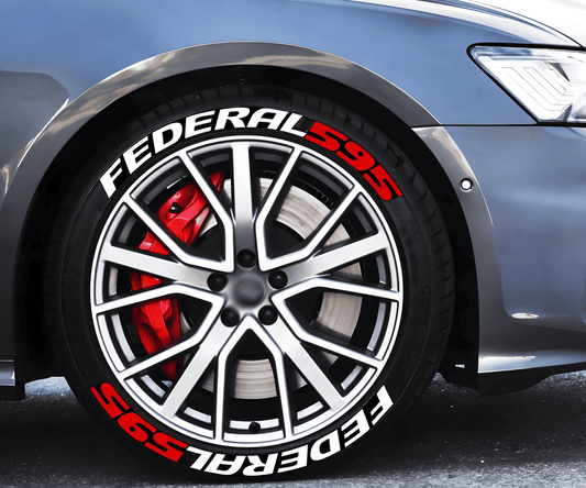 A close-up of a silver sports car's front wheel showcasing ChroMorpher FEDERAL 595 3D tire lettering decals in bold white and red. The wheel features a multi-spoke alloy rim with a red performance brake caliper and a ventilated brake disc visible behind it. The high-performance tire, enhanced by the lettering, gives the vehicle an aggressive and race-inspired look. The car is parked on an asphalt surface with a blurred background.