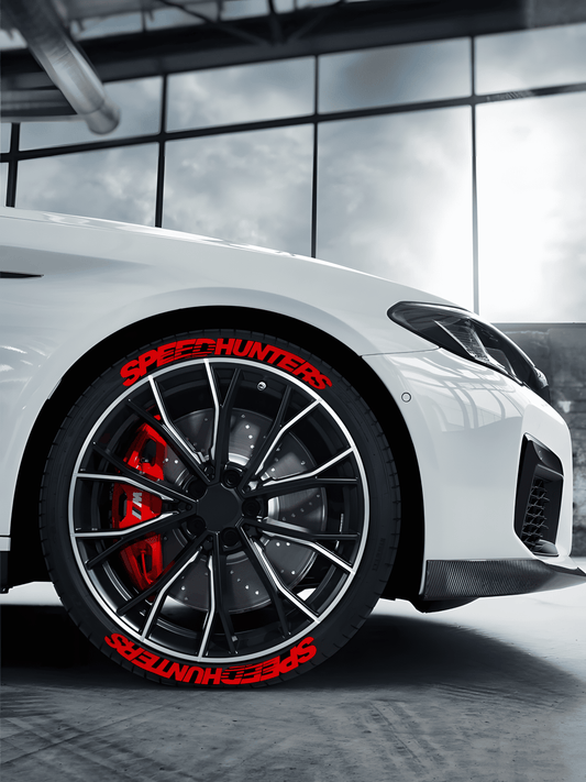 A white sports car featuring ChroMorpher SpeedHunters Red 3D Tire Lettering Decals on its black tires. The bold red SpeedHunters text enhances the aggressive racing aesthetic, complementing the red brake calipers and black multi-spoke wheels for a striking performance look.