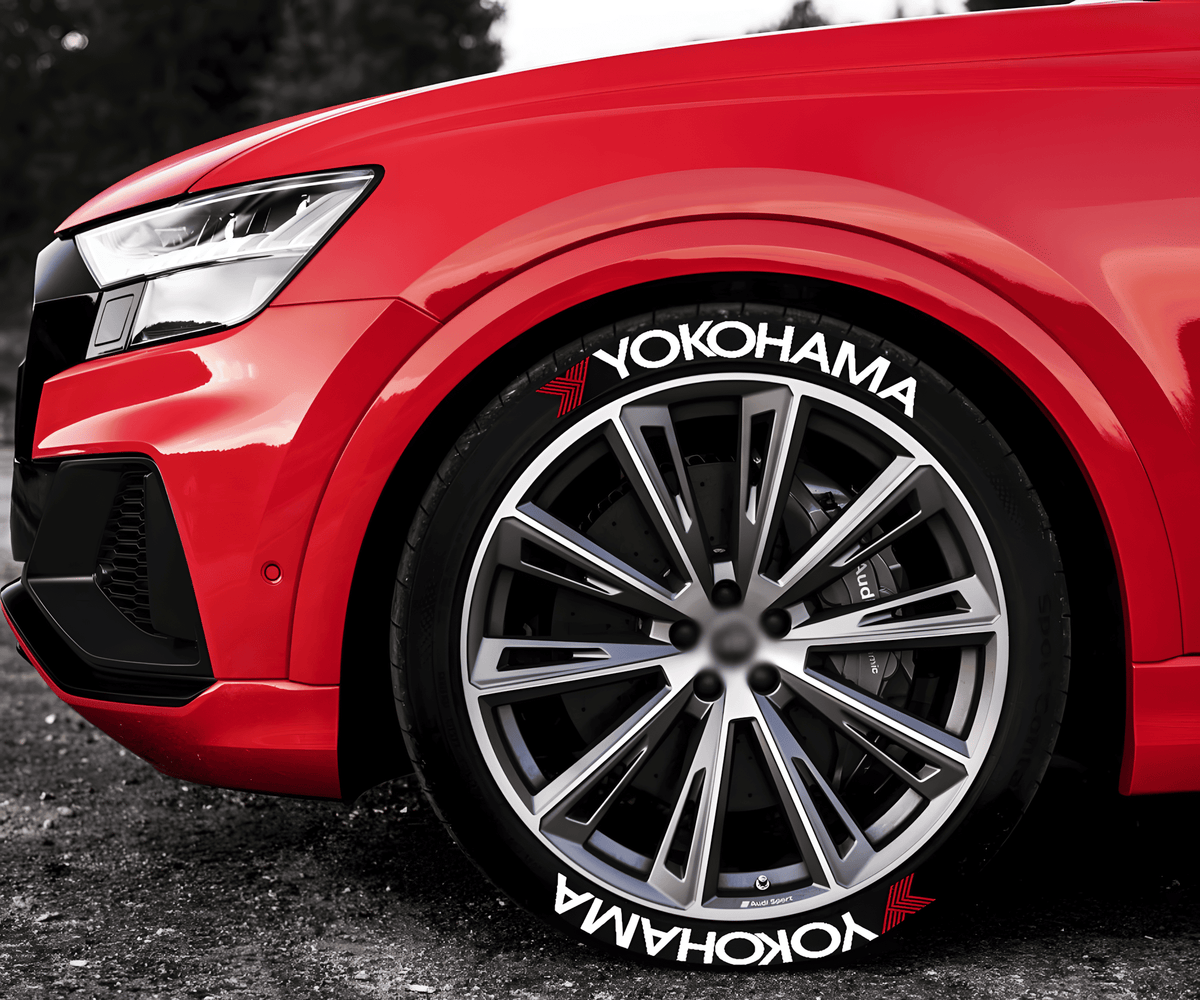 Close-up of a red sports car with Yokohama 3D tire lettering decals on large multi-spoke alloy wheels. The white Yokohama branding with red accents enhances the vehicle's aggressive and high-performance aesthetic, perfect for motorsport enthusiasts and street racers.