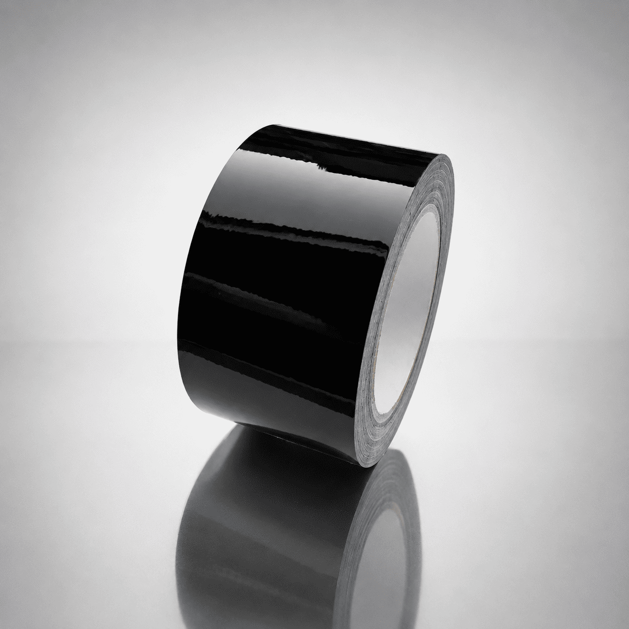A roll of glossy black tape standing upright on a reflective light gray surface, with a soft reflection below.