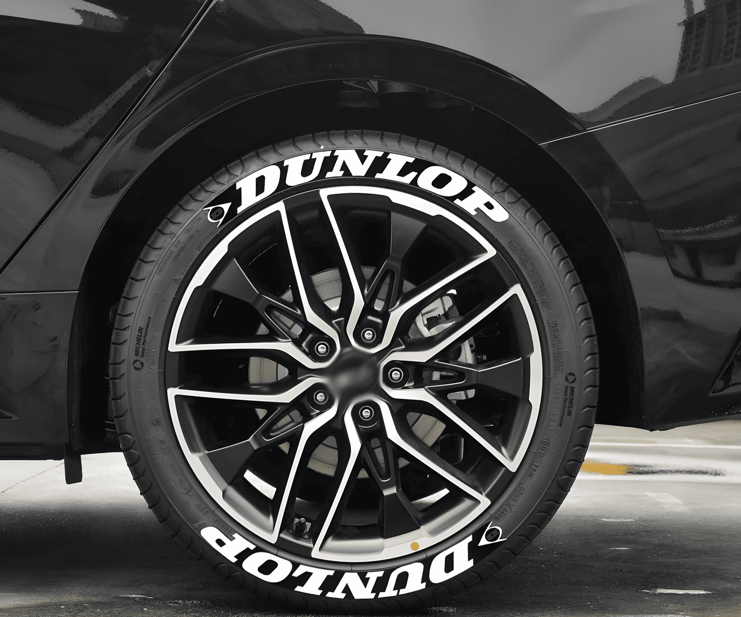 A black sports car fitted with ChroMorpher Dunlop Racing Tire Lettering Decals. The bold white "DUNLOP" text with a sleek racing-inspired design enhances the tire’s sporty look, perfect for high-performance vehicles.