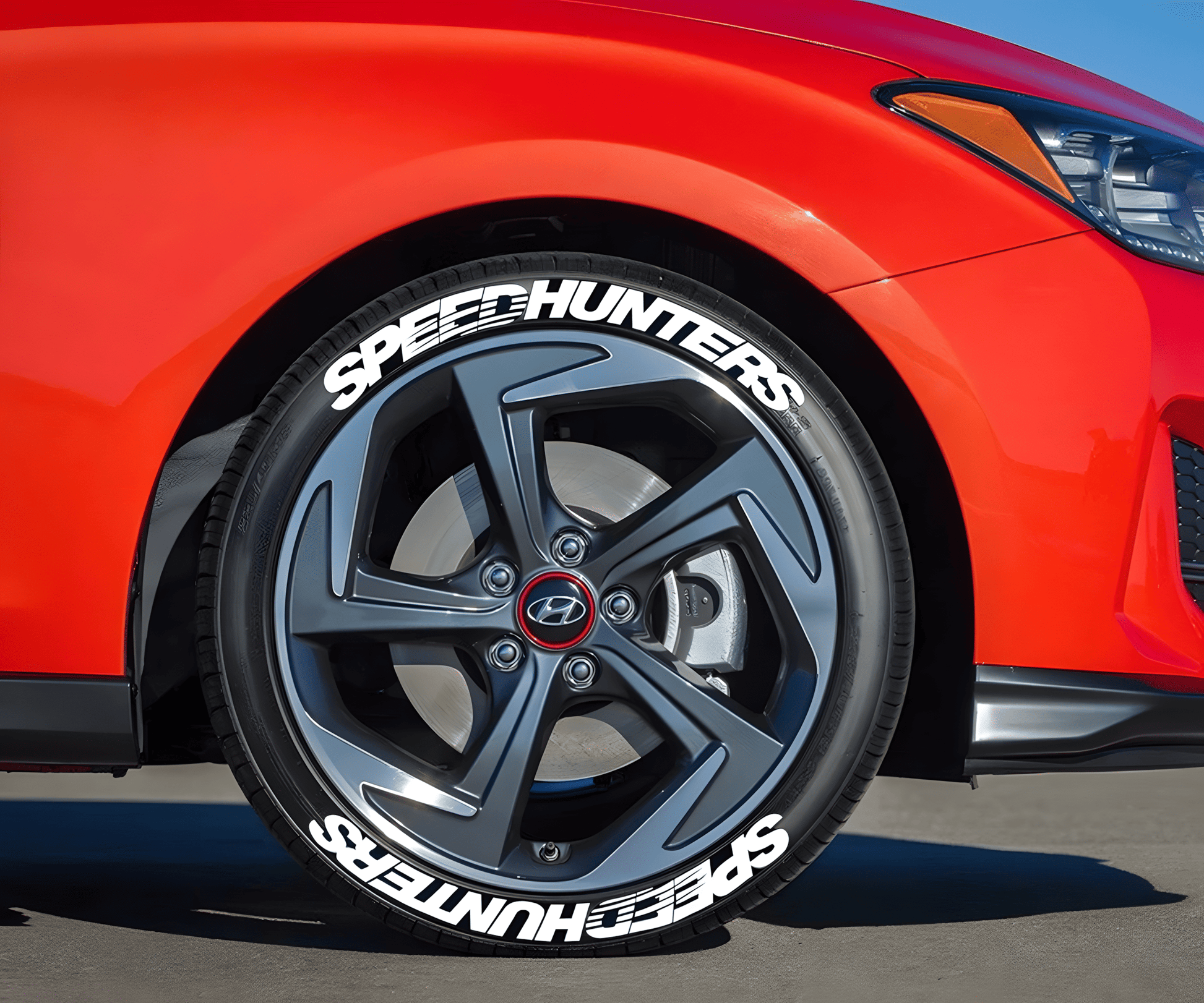 A close-up of a red Hyundai with ChroMorpher SpeedHunters 3D Tire Lettering Decals applied to the tires. The white SpeedHunters lettering adds a bold racing-inspired look, enhancing the vehicle's sporty appeal. The decals are precisely positioned for a professional and aggressive aesthetic.