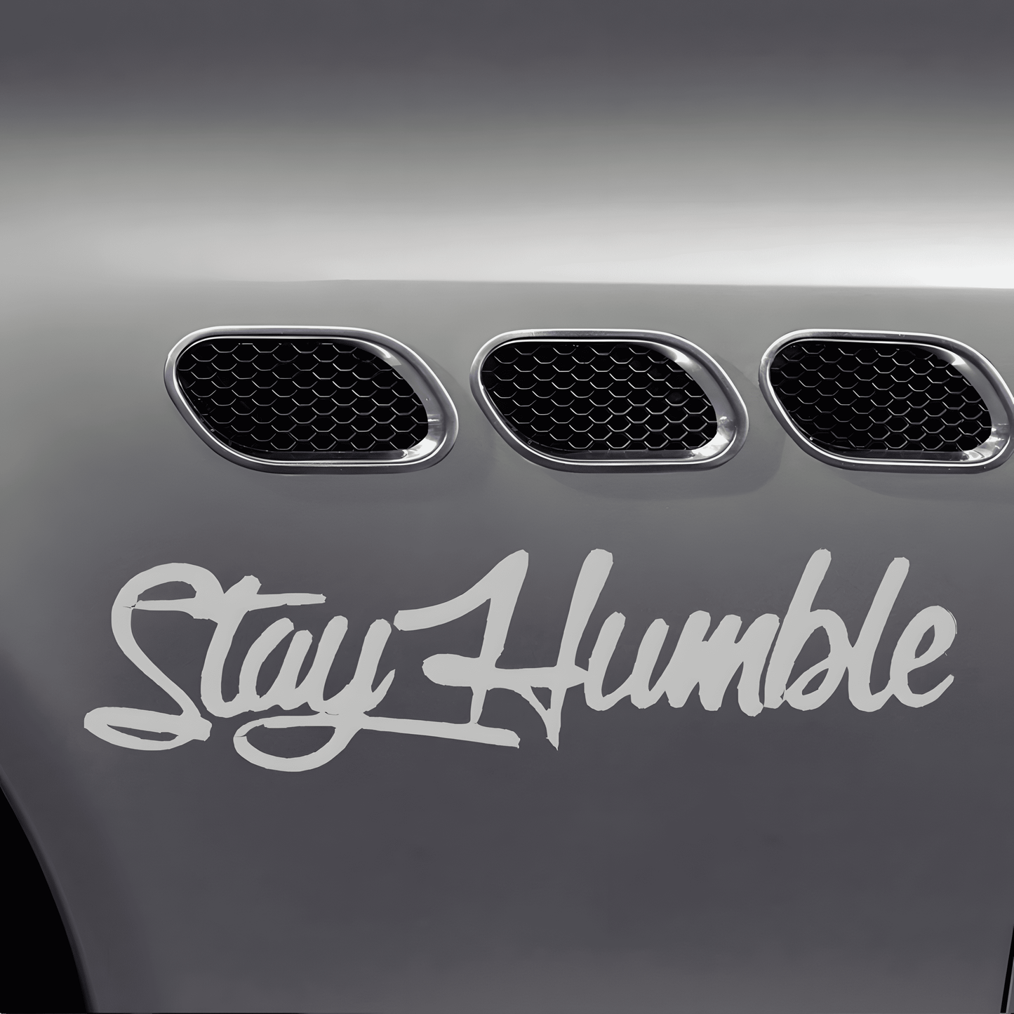 A silver-white "Stay Humble" car sticker applied to a vehicle fender, featuring a glossy finish and bold cursive font. This waterproof, self-adhesive decal is perfect for car bodies, windows, bumpers, and other smooth surfaces, adding a stylish and motivational statement.