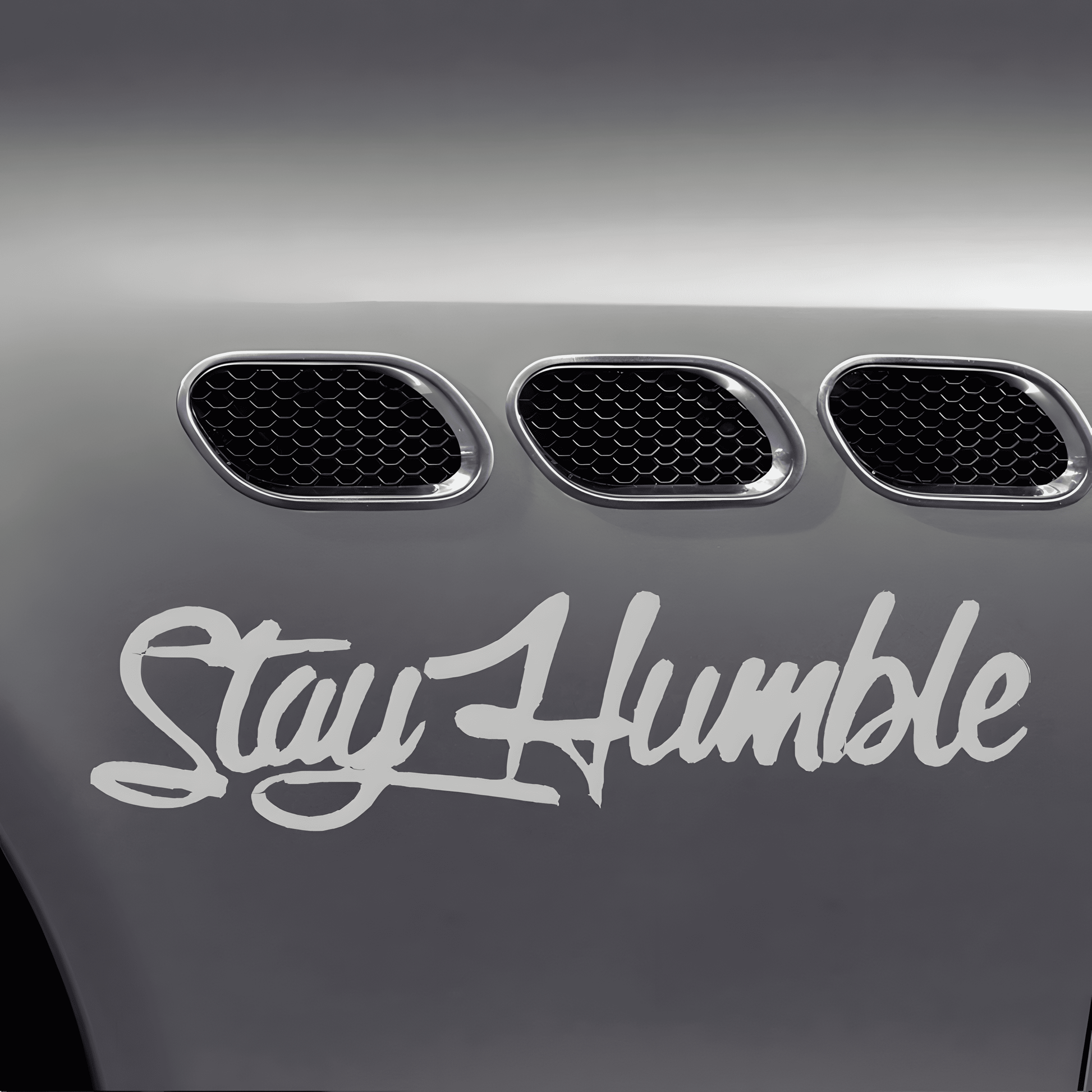 A silver-white "Stay Humble" car sticker applied to a vehicle fender, featuring a glossy finish and bold cursive font. This waterproof, self-adhesive decal is perfect for car bodies, windows, bumpers, and other smooth surfaces, adding a stylish and motivational statement.