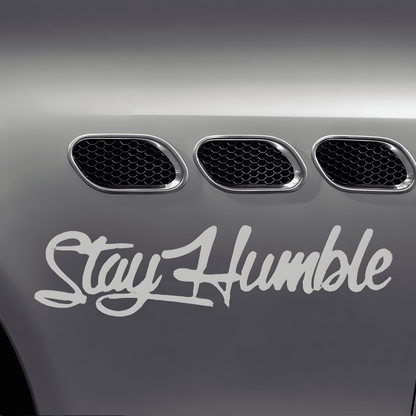 A silver-white "Stay Humble" car sticker applied to a vehicle fender, featuring a glossy finish and bold cursive font. This waterproof, self-adhesive decal is perfect for car bodies, windows, bumpers, and other smooth surfaces, adding a stylish and motivational statement.