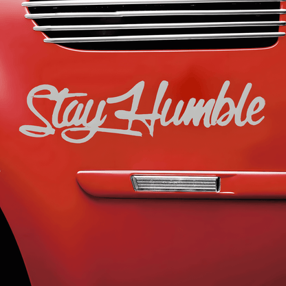 A silver-white "Stay Humble" car sticker applied to a red vehicle, featuring a glossy finish and stylish cursive font. This waterproof, self-adhesive decal is designed for car bodies, windows, bumpers, and other smooth surfaces, offering a sleek, motivational aesthetic.