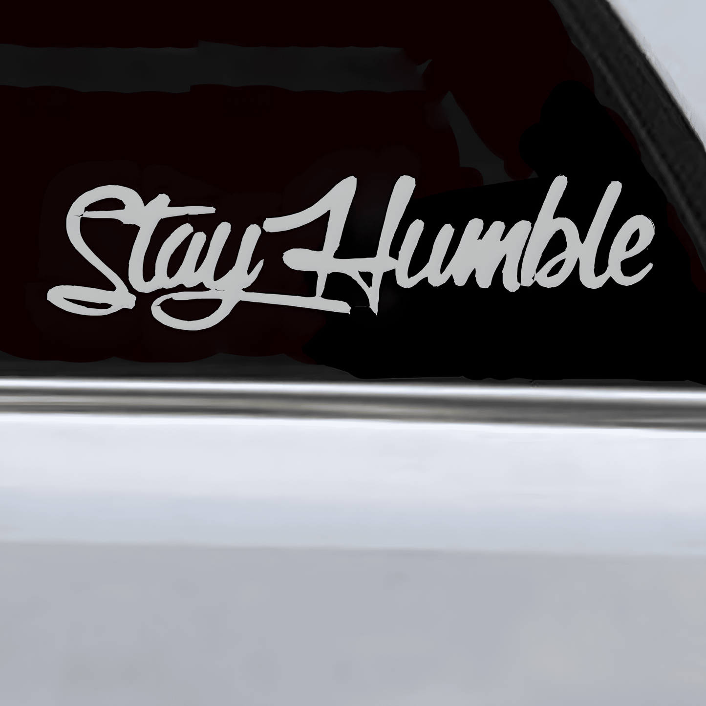 A silver-white "Stay Humble" car sticker applied to a vehicle window, featuring a glossy finish and stylish cursive font. This waterproof, self-adhesive decal enhances car windows, bumpers, and other smooth surfaces, adding a motivational and sleek touch.