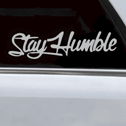 A silver-white "Stay Humble" car sticker applied to a vehicle window, featuring a glossy finish and stylish cursive font. This waterproof, self-adhesive decal enhances car windows, bumpers, and other smooth surfaces, adding a motivational and sleek touch.
