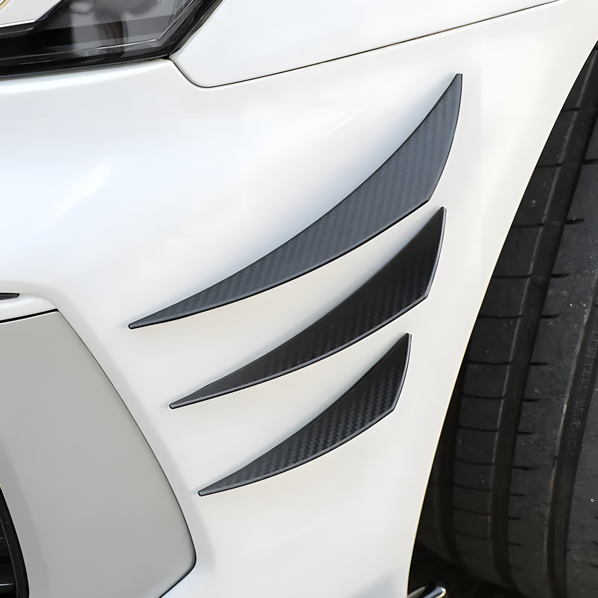 Close-up of a white car’s front bumper featuring universal carbon fiber-style canard fins, designed to enhance aerodynamics and provide a sporty, aggressive look. These lightweight, easy-to-install bumper fins help reduce drag and improve stability.