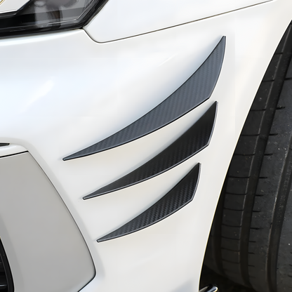 Close-up of a white car’s front bumper featuring universal carbon fiber-style canard fins, designed to enhance aerodynamics and provide a sporty, aggressive look. These lightweight, easy-to-install bumper fins help reduce drag and improve stability.