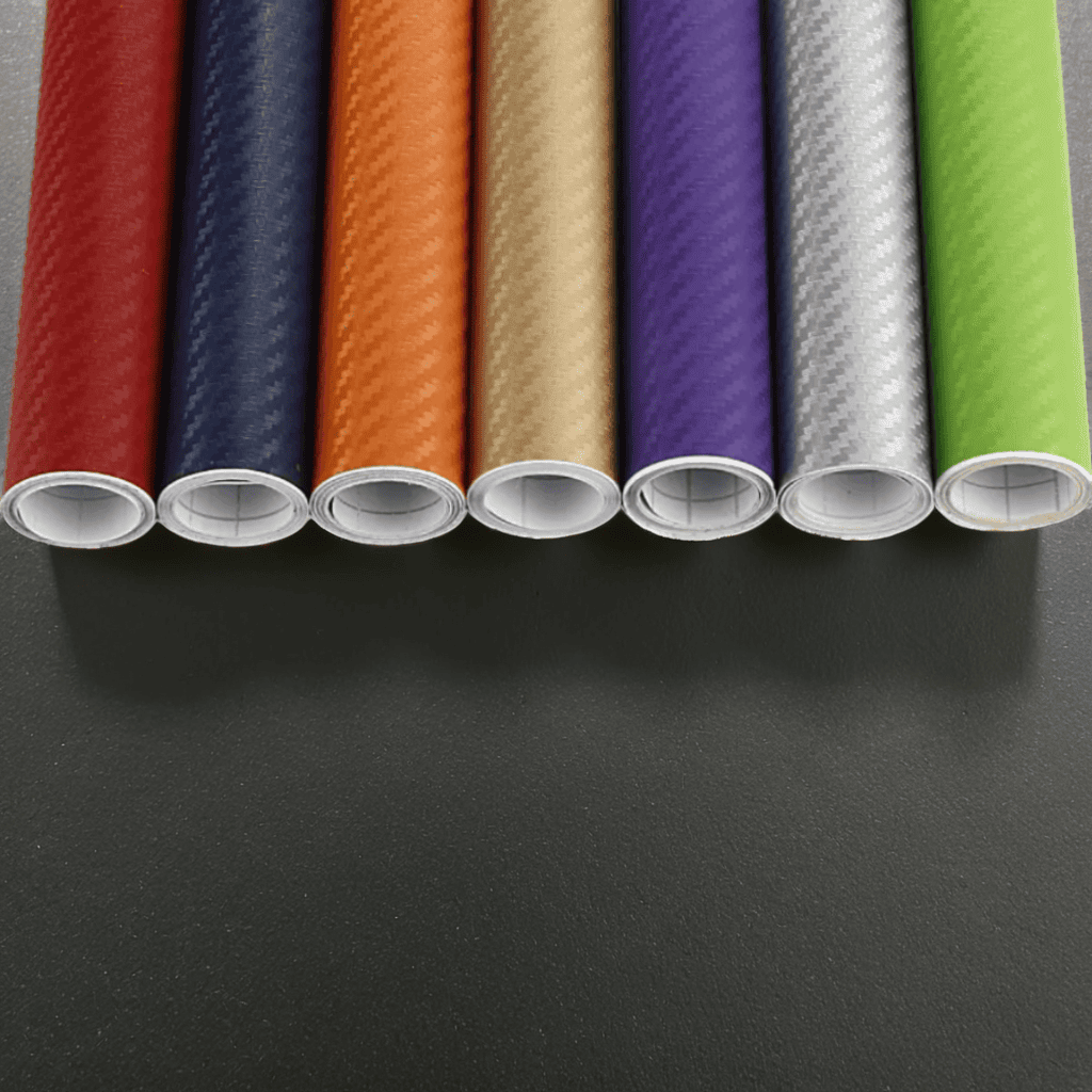 Rolls of ChroMorpher Wide Carbon Vinyls in vibrant colors including red, blue, orange, gold, purple, silver, and green, showcasing a realistic 3D carbon fiber texture for vehicle and surface customization.