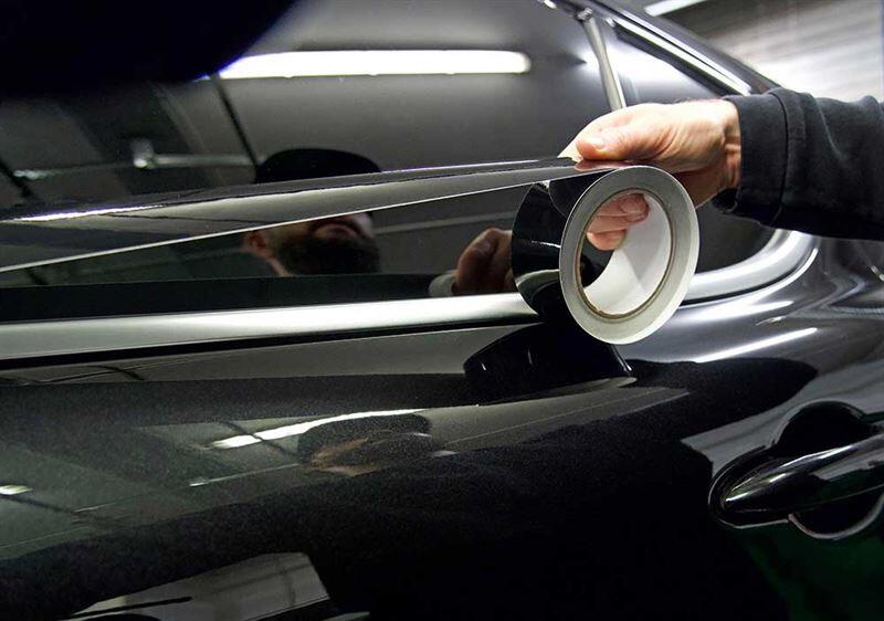 A person applying black vinyl tape to the chrome trim of a car window for a chrome delete modification.