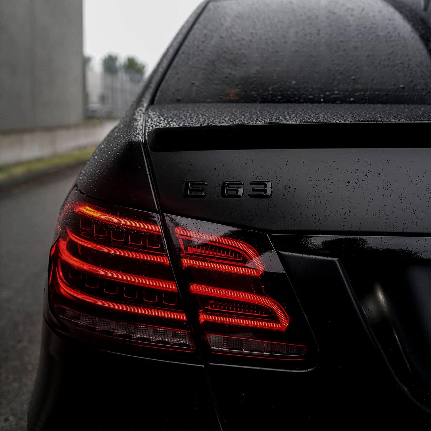 Rear view of a Mercedes-Benz E63 with chrome delete treatment, featuring sleek, glossy black finish and red taillights in a rainy urban setting.